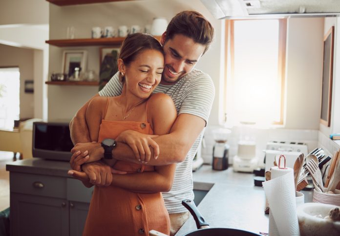 Love, romance and fun couple hugging, cooking in a kitchen and sharing an intimate moment. Romantic boyfriend and girlfriend embracing, enjoying their relationship and being carefree together